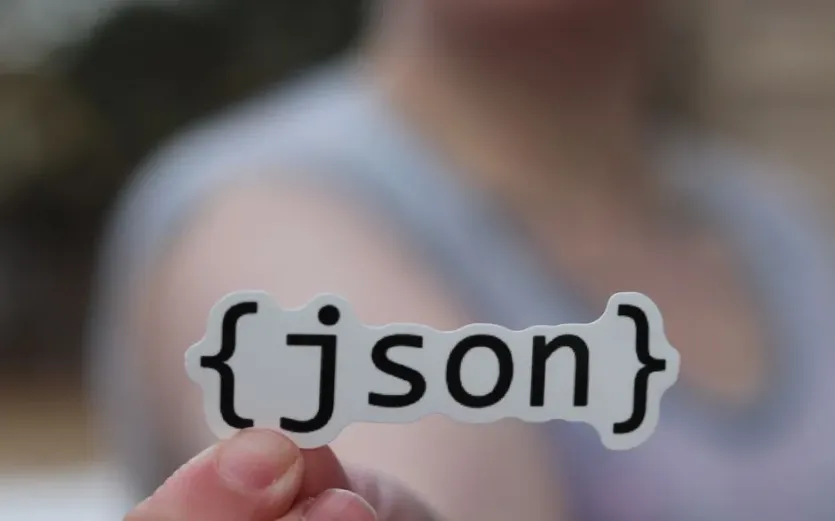 노트북 앞에서 중괄호로 감싼 json 이라는 글자가 적힌 스티커를 들어 보이는 손